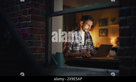 Focused Man Working on Laptop Late in the Evening Finishing Important Project at Home Office. Freelancer Working Remotely. Warm Lit Cozy Apartment. View From Outdoors into Window. Stock Photo