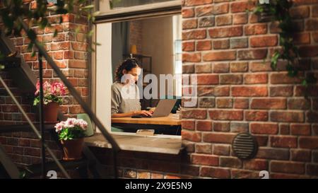 Hispanic Woman Using Laptop Computer, Sitting in Living Room im Her Apartment. Professional Female Working Remotely Online, Planning, Brainstorming New Project. Shot Into the Window From Outside. Stock Photo