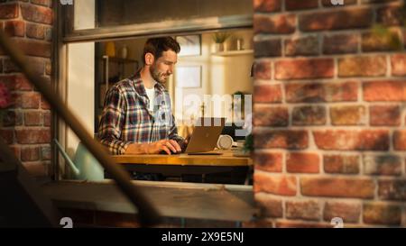Male Software Engineer Coding on Laptop Computer, Working in Modern Apartment. Professional IT Specialist Designing Application. Freelancer Doing Remotre Work. View From Outdoors into Window. Stock Photo