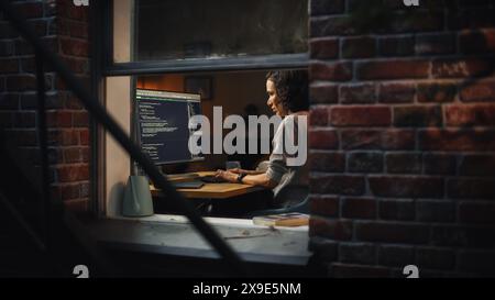 Night Home Office: Young Hispanic Woman Working on Desktop Computer. Software Developer, e-Commerce Entrepreneur Writing Code for e-Business App, Designing Program. View From Outdoors into Window. Stock Photo