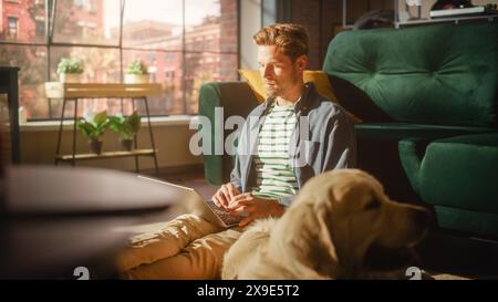 Handsome Young Man Using Laptop, Working from Home Living Room. Male Sitting on Floor, Using Computer, Golden Retriever Dog Sitting Next to Him and Waiting to Go For a Walk. Stock Photo