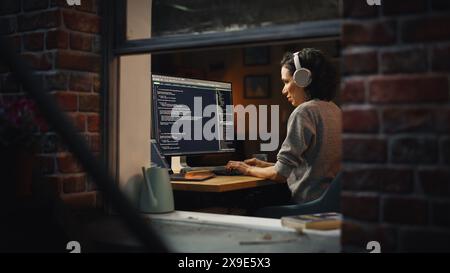 Hispanic Female Programmer in Headphones Listening Podcast and Working on Desktop Computer, Screen Shows Coding Language User Interface. Digital Entrepreneur Creating Modern Software. Stock Photo