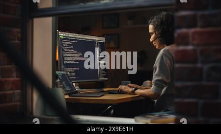 Night Home Office: Young Hispanic Woman is Working on Desktop Computer. Software Developer, e-Commerce Entrepreneur Writing Code for e-Business App, Designing Program. View From Outdoors into Window. Stock Photo