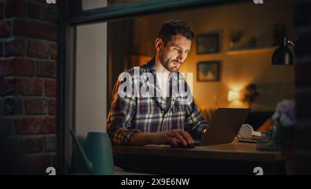 Serious Man Working on Laptop Late in the Evening Finishing Important Project at Home Office. Freelancer Working Remotely. Warm Lit Cozy Apartment. View From Outdoors into Window. Stock Photo