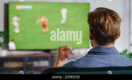 Sports Fan Watches Important Soccer Match on TV at Home, He Aggressively Gestures with the Fist, Cheering for His Team. Cozy Living Room in Modern Stylish Loft Apartment. Stock Photo