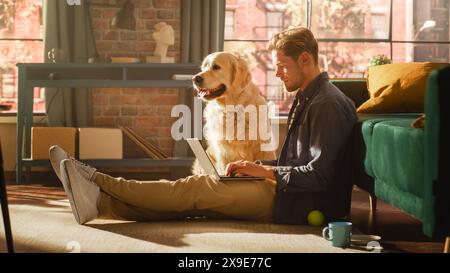 Portrait of a Handsome Young Adult Male Sitting on a Floor and Using Laptop Computer in Sunny Loft Living Room. Golden Retriever Dog Sitting Next to Him and Wanting to Play and Get Petted. Stock Photo