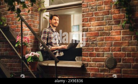 Handsome Creative Male Works on a Laptop Computer and Sitting on a Windowsill of his Cozy Apartment. Professional Specialist Working Remotely from Home. Shot Through Building Window. Stock Photo