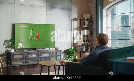 Sports Fan Watches Important Soccer Match on TV at Home, He Is Focused on Intense Championship, Cheering for His Team. His Successful Team Scores and Leading the Game. Stock Photo