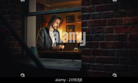 Professional Male Programmer Writing Code for Complex Project and Working Remotely on Laptop From Home Office. Freelance Software Engineer Designs App From Apartment. View From Outdoors into Window. Stock Photo