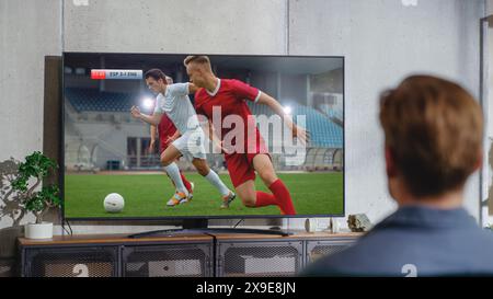 Sports Fan Watches Important Soccer Match on TV at Home, He Is Focused on Intense Championship, Cheering for His Team. His Favourite Football Team Scores and Leading the Game. Stock Photo