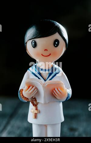 a figurine of a first communion boy, wearing a white sailor suit, a rosary and a bible, standing on a rustic gray wooden surface Stock Photo