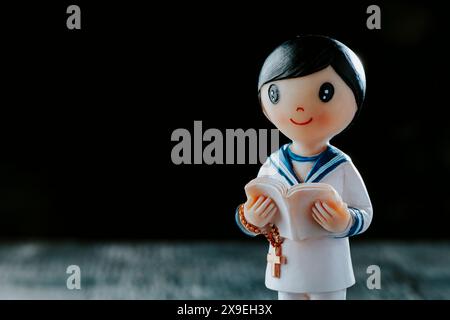 closeup of a figurine of a first communion boy, wearing a white sailor suit, a rosary and a bible, standing against a black background with some blank Stock Photo