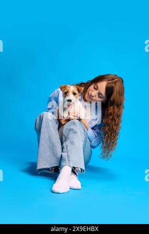 Portrait of young woman posing with her Jack Russel dog against vibrant blue studio background, enjoying time with pet. Stock Photo