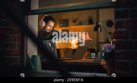 Professional Software Developer Stretches and Continues Typing on Laptop, Writing Code and Working on the Project. Remote Work at Night Time Concept. View From Outdoors into Window. Stock Photo