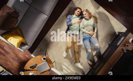Top View Apartment: Beautiful Couple Relaxing on a Carpet in the Living Room, Using Smartphone. Girlfriend and Boyfriend Enjoy being Together, Online Shopping, e-business, Surfing the Internet. Stock Photo