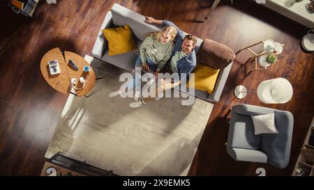 Top View Apartment: Happy Couple In Love Using Laptop in Stylish Living Room, Hug and Look Up at Camera. On Sofa: Girlfriend With Her Boyfriend Use Portable Computer for Online Shopping, e-business. Stock Photo