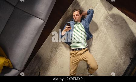 Apartment: Handsome Man Lying on a Living Room Floor, Using Smartphone. Guy Relaxes on the Carpet. Freelancer Does Remote Work, E-business, Online Shopping, Social Media Browsing. Top View. Stock Photo