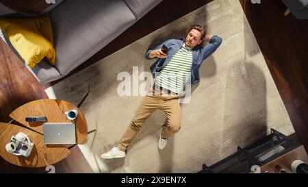Apartment: Handsome Man Lying on a Living Room Floor, Using Smartphone. Guy Relaxes on Carpet. Freelancer Does Remote Work, E-business, Online Shopping, Social Media Browsing. Top View. Stock Photo