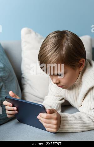 Boy spends time behind a screen on a tablet on the sofa in the living room. Digital device. Technologies. Stock Photo