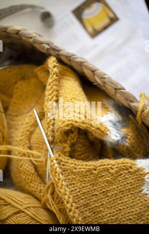 Close-up photography of yellow alpaca wool knitted sweater sleeves in progress with magic loop technique in a basket with yarn balls and printed patte Stock Photo