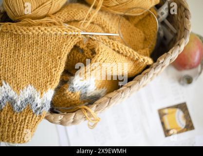 Close-up photography of yellow alpaca wool knitted sweater sleeves in progress with magic loop technique in a basket with yarn balls and printed patte Stock Photo