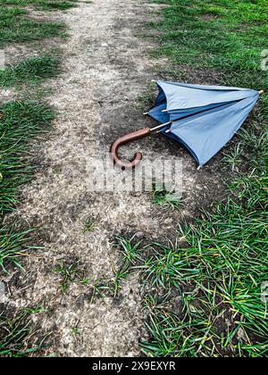 Grey umbrella with a wooden handle discarded on a dry muddy footpath surrounded by grass Stock Photo