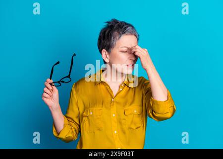 Photo of tired upset lady dressed shirt holding eyewear having head ache isolated blue color background Stock Photo