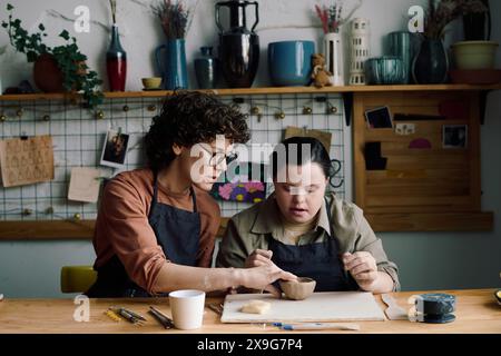 Mature female potter sitting at table teaching young woman with Down syndrome how to make ceramic vessel, copy space Stock Photo