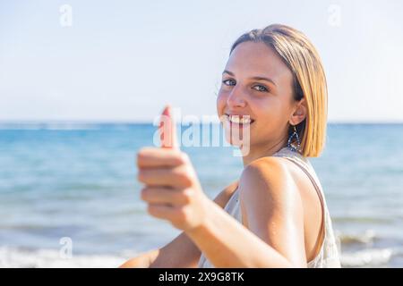 A redhead young woman is smiling and giving a thumbs up while standing on a beach Stock Photo