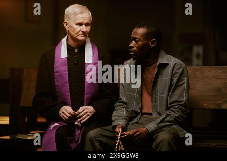 African American man holding rosary sitting on pew having conversation with senior Caucasian Catholic priest, medium shot Stock Photo