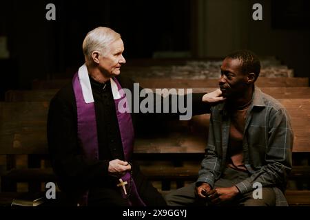 Medium shot of senior Caucasian Catholic priest and young African American man sitting on pew in church having conversation Stock Photo