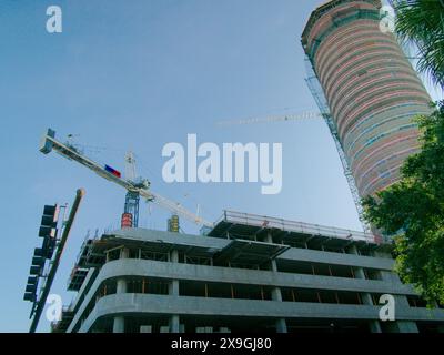 View up through green trees in foreground to multi-story parking garage, building and cranes under construction. Blue sky and fencing. No people Stock Photo