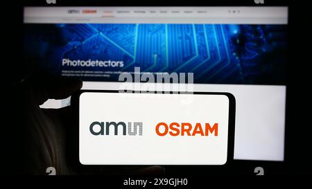 Person holding cellphone with logo of Austrian electronics company ams OSRAM AG in front of business webpage. Focus on phone display. Stock Photo