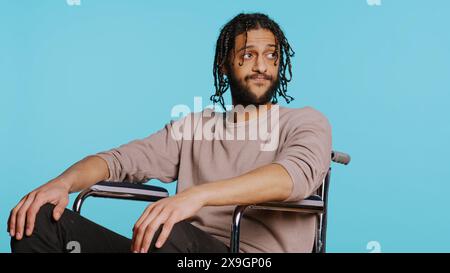 Bored Middle Eastern man with disability in wheelchair impatiently tapping fingers on knee, waiting. Annoyed person with paraplegia looking around, isolated over studio background, camera B Stock Photo