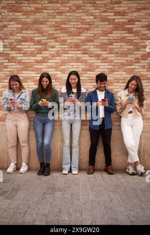 Multiracial smiling group young generation z leaning on brick wall using phones outdoors. Stock Photo