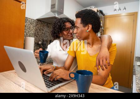 Romantic latin gay couple using laptop and drinking coffee sitting in a domestic kitchen at home Stock Photo