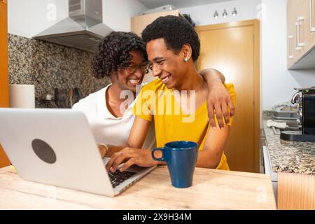 Gay couple laughing and embracing while using laptop drinking coffee sitting in the kitchen at home Stock Photo