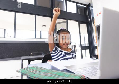 In school, young biracial girl wearing headphones raising hand in the classroom Stock Photo