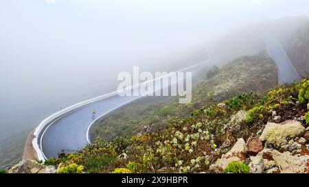 Foggy mountain road with a lone cyclist winding through lush green scenery Stock Photo