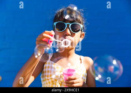 Biracial girl wearing sunglasses and a sundress enjoys blowing bubbles outdoors Stock Photo