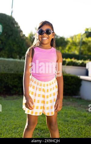 Young biracial girl in sunglasses, pink top, smiling brightly on grass outdoors Stock Photo