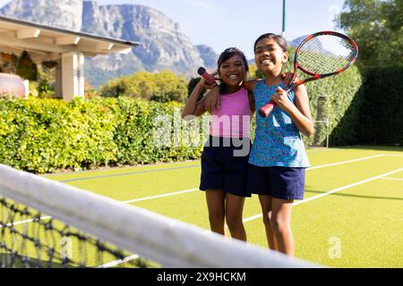 Outdoors, two biracial young sisters smile and hold tennis rackets on tennis court Stock Photo