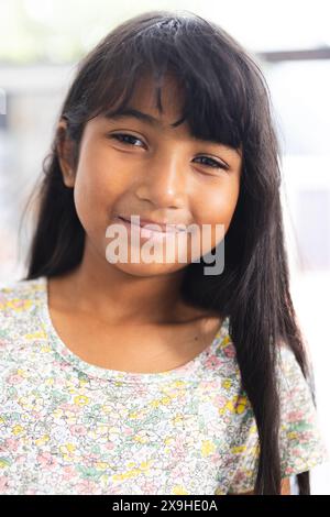 Biracial girl with a warm smile, wearing a floral dress in a school classroom Stock Photo