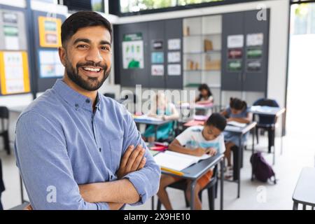 Young Asian male teacher with a beard stands confidently in a school classroom with copy space. He is a teacher, with students focused on their work i Stock Photo