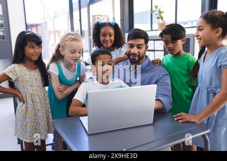 In school, young Asian male teacher and diverse students gather around a laptop in the classroom Stock Photo