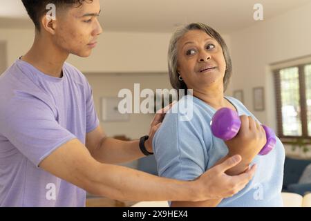 At home, biracial male physiotherapist and senior female patient doing arm exercises, using dumbbells . They are in bright, modern living room with la Stock Photo
