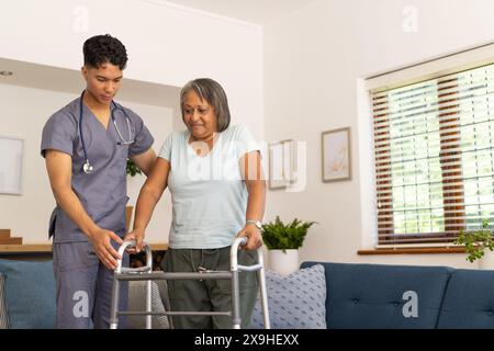 At home, young biracial male nurse assisting senior female patient. They are in bright, cozy living room with a blue background, sofa, and large windo Stock Photo