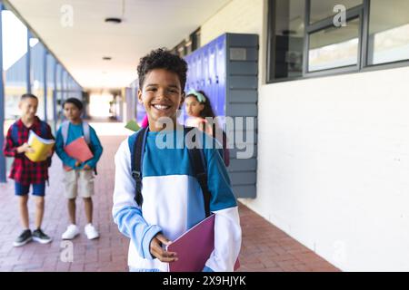 Biracial boy with a bright smile stands in a school hallway, holding a folder. Behind him, two biracial boys and two biracial girls carry backpacks, s Stock Photo