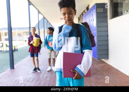 Biracial boy in blue stands thoughtfully in a school corridor, holding a book Stock Photo