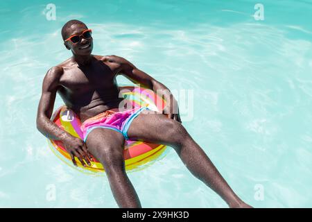An African American young male, wearing sunglasses, floating outside on pool ring. Showcasing his fit physique and dark skin, smiling in sunlight, una Stock Photo